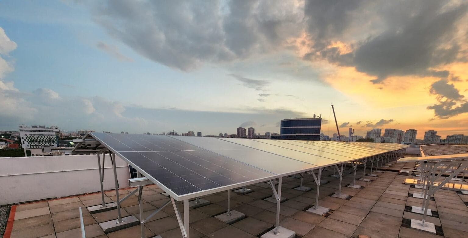 A long run of solar photovoltaic panels on the flat, paved rooftop of the Singapore PharmaTech facility, seen end-on down the row at sunset. The modules are tilted on slim aluminium A-frame supports, each leg footed on its own square concrete ballast block; the nearest panels read dark blue with the grid of cells visible, while those further along mirror the sky. Past the white parapet wall on the left, the surrounding skyline carries HDB blocks, a blue-glass office building and a red construction crane, under heavy cloud that breaks into warm orange light on the right.